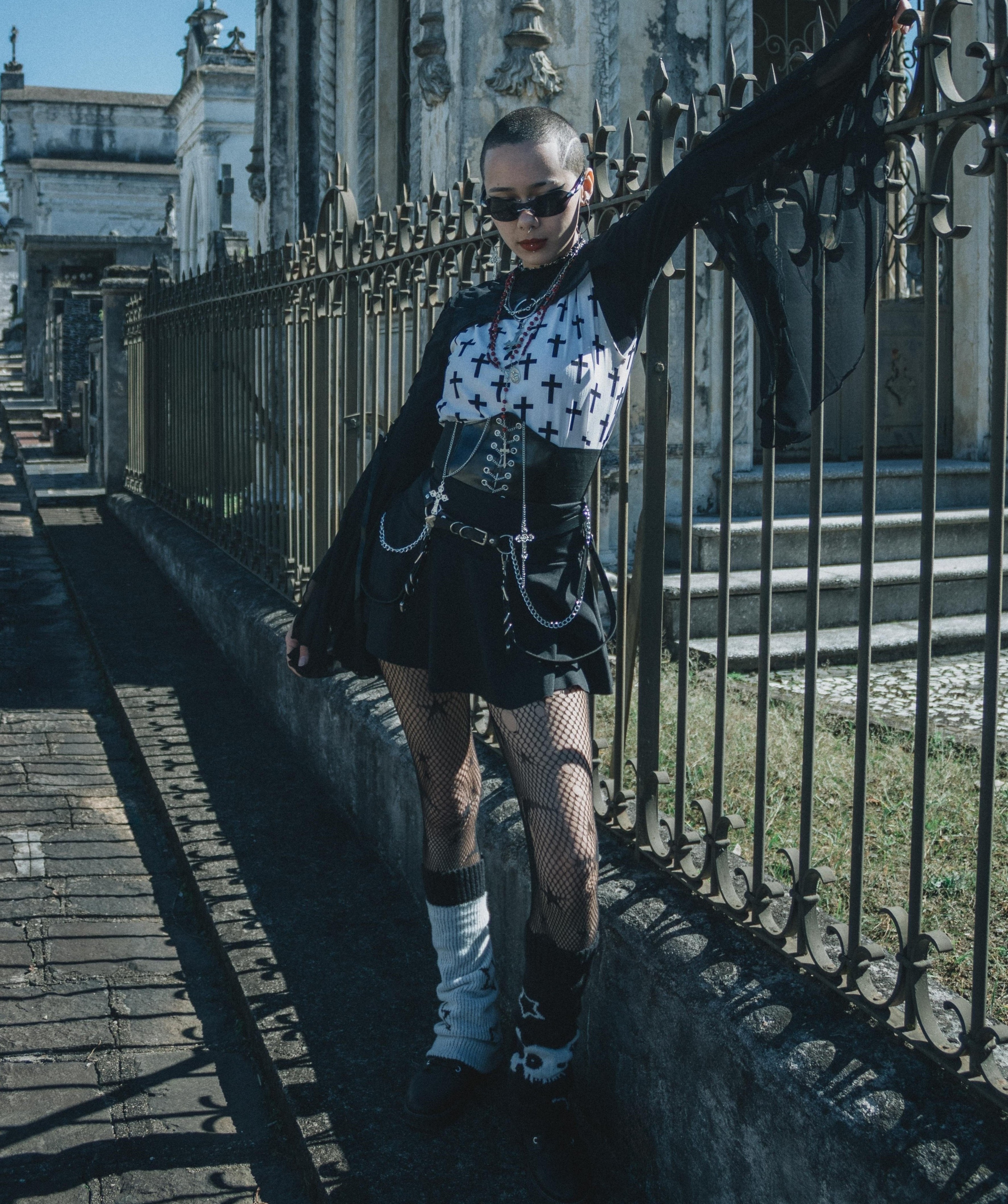 Person wearing a white cross-print top over a black long-sleeve layer, 
          a black skirt, fishnet tights, layered jewelry, and mismatched leg warmers.