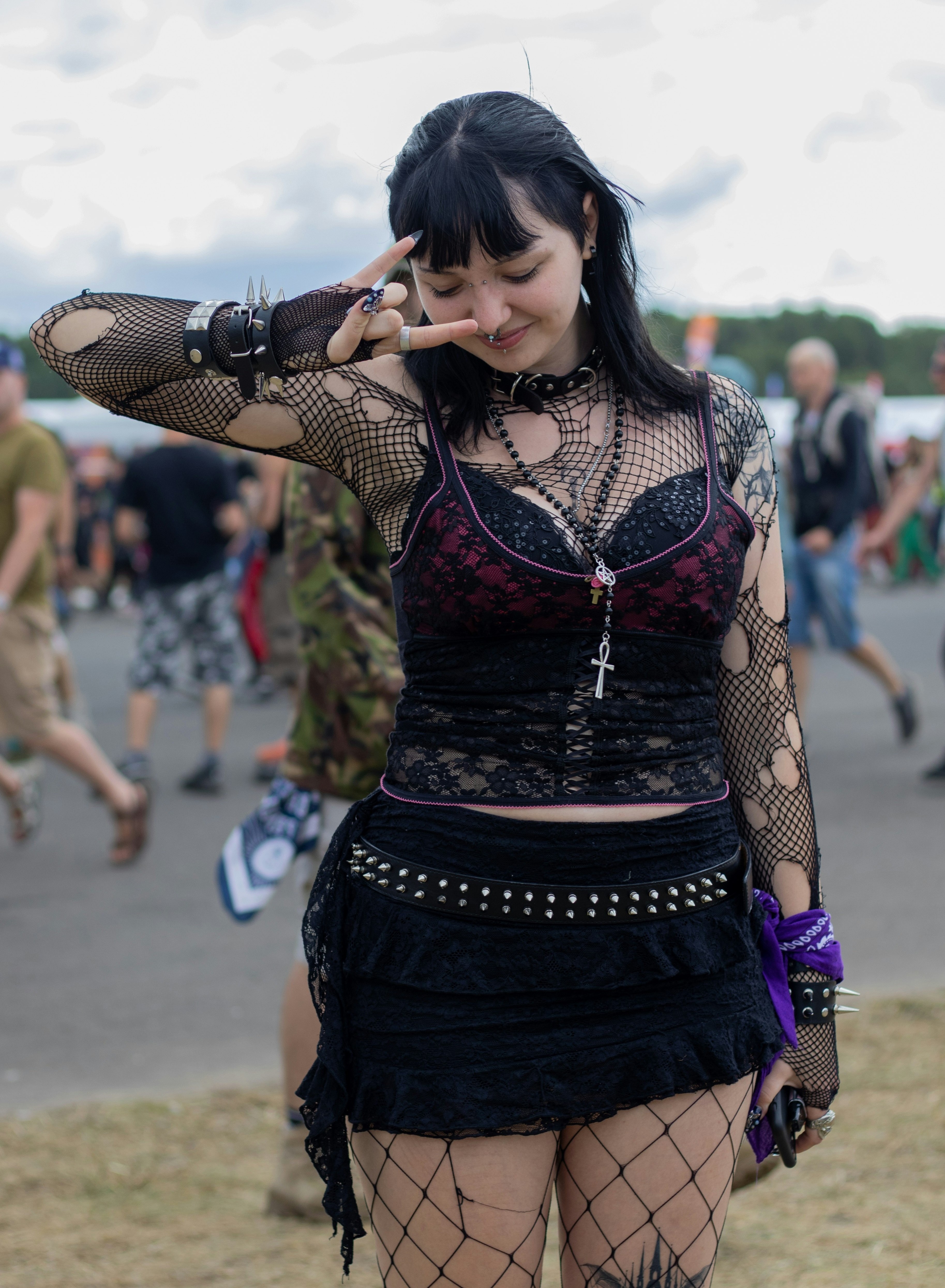 Person wearing a black lace top with pink trim, ripped fishnet sleeves, layered necklaces, a studded belt, black skirt, and fishnet tights.