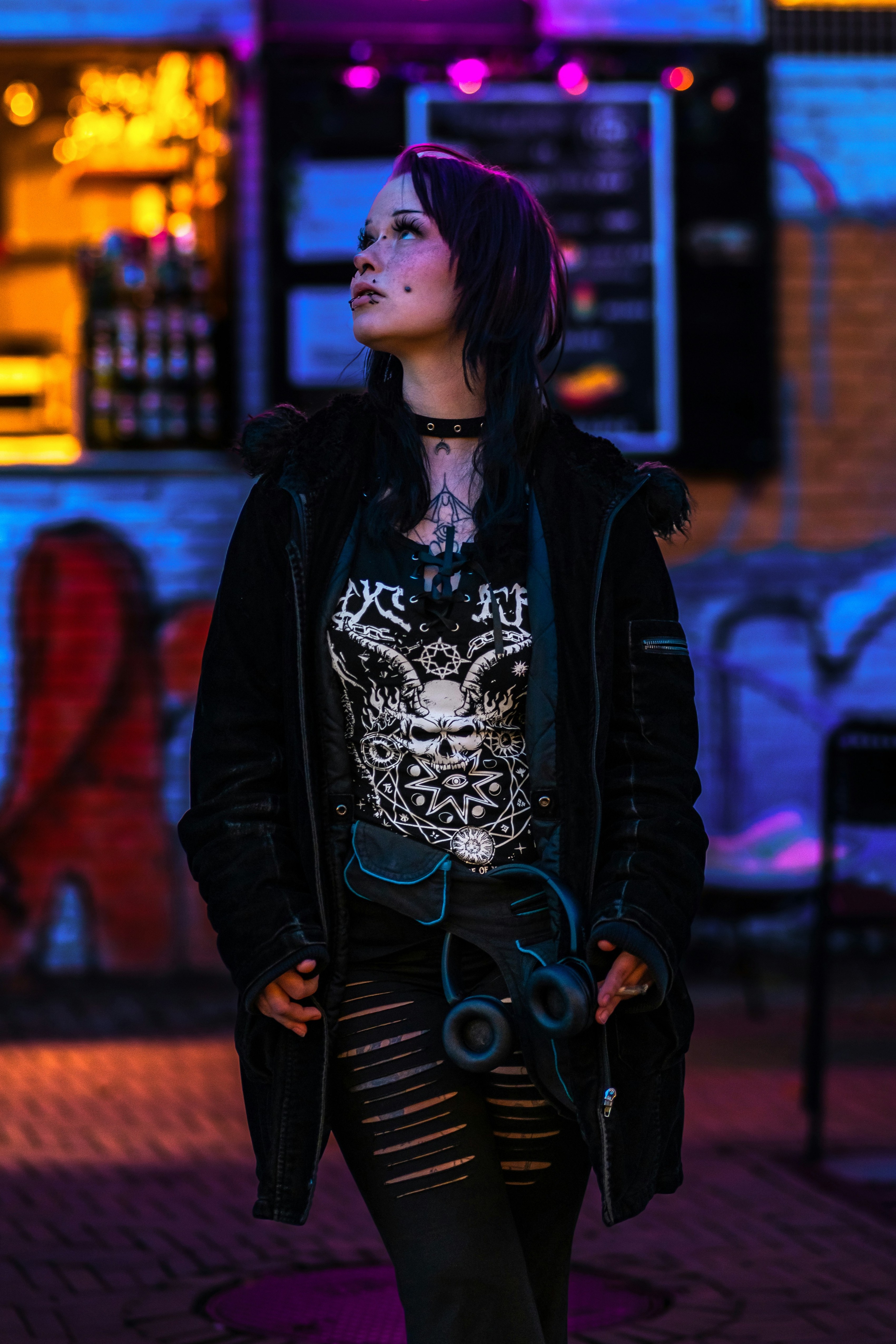 Person wearing a black graphic top, black jacket, ripped black pants, headphones, and a choker in colorful neon lighting.
