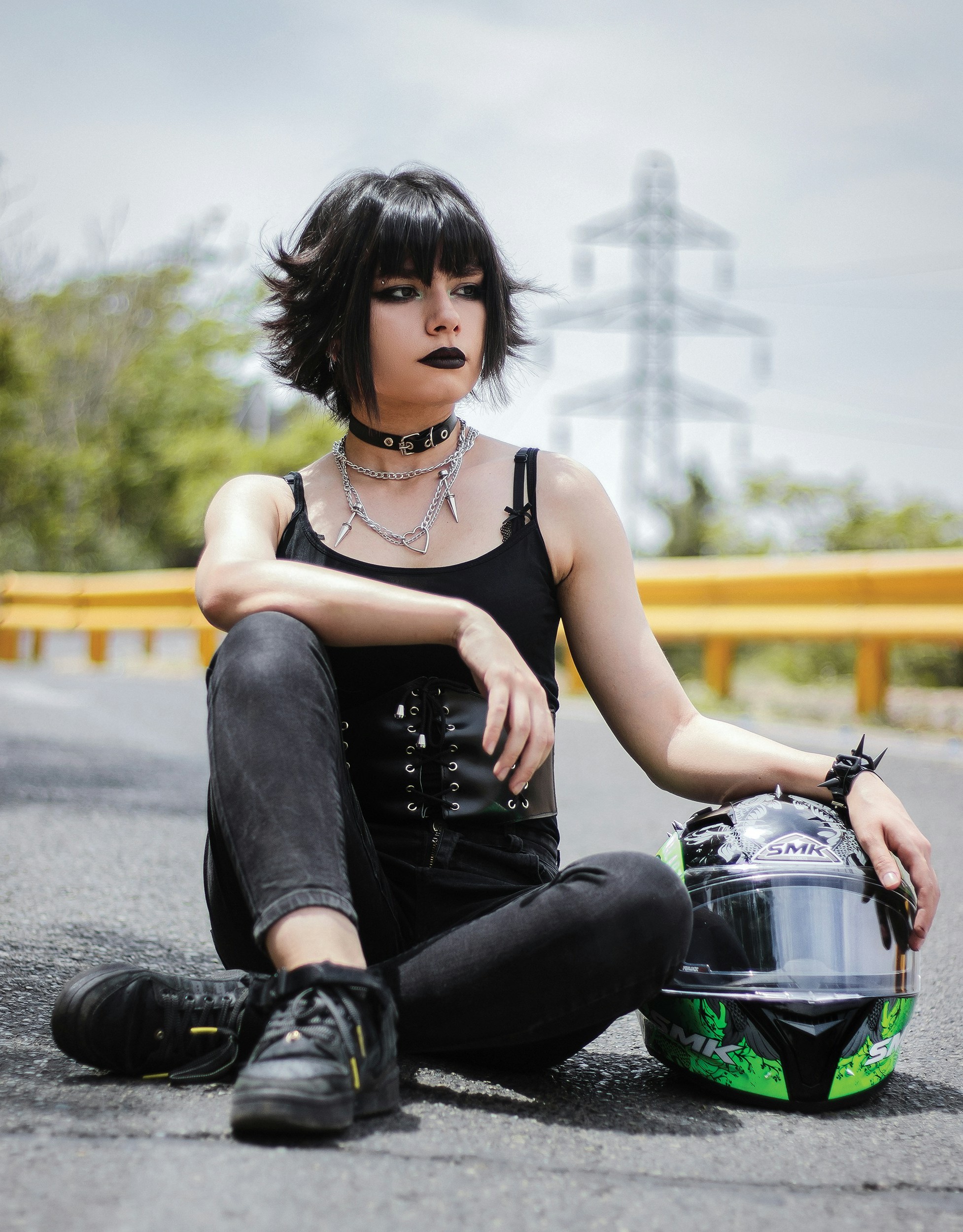 Person sitting on the road wearing a black tank top, corset-style belt, layered necklaces, spiked bracelet, black pants, and black shoes.
