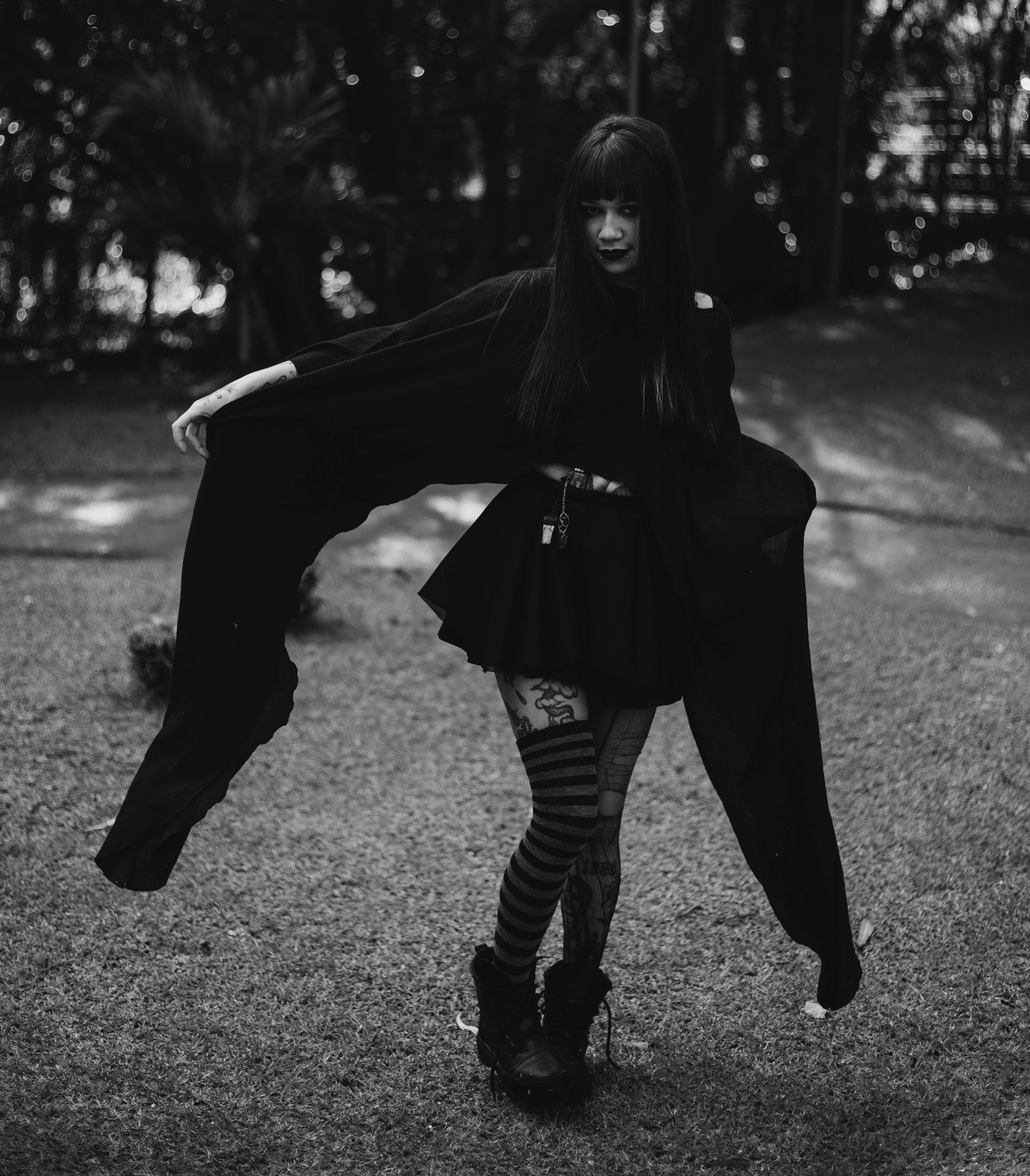 Black and white image of a person wearing a flowing black outfit with wide sleeves, a short skirt, patterned tights, striped socks, and black boots.