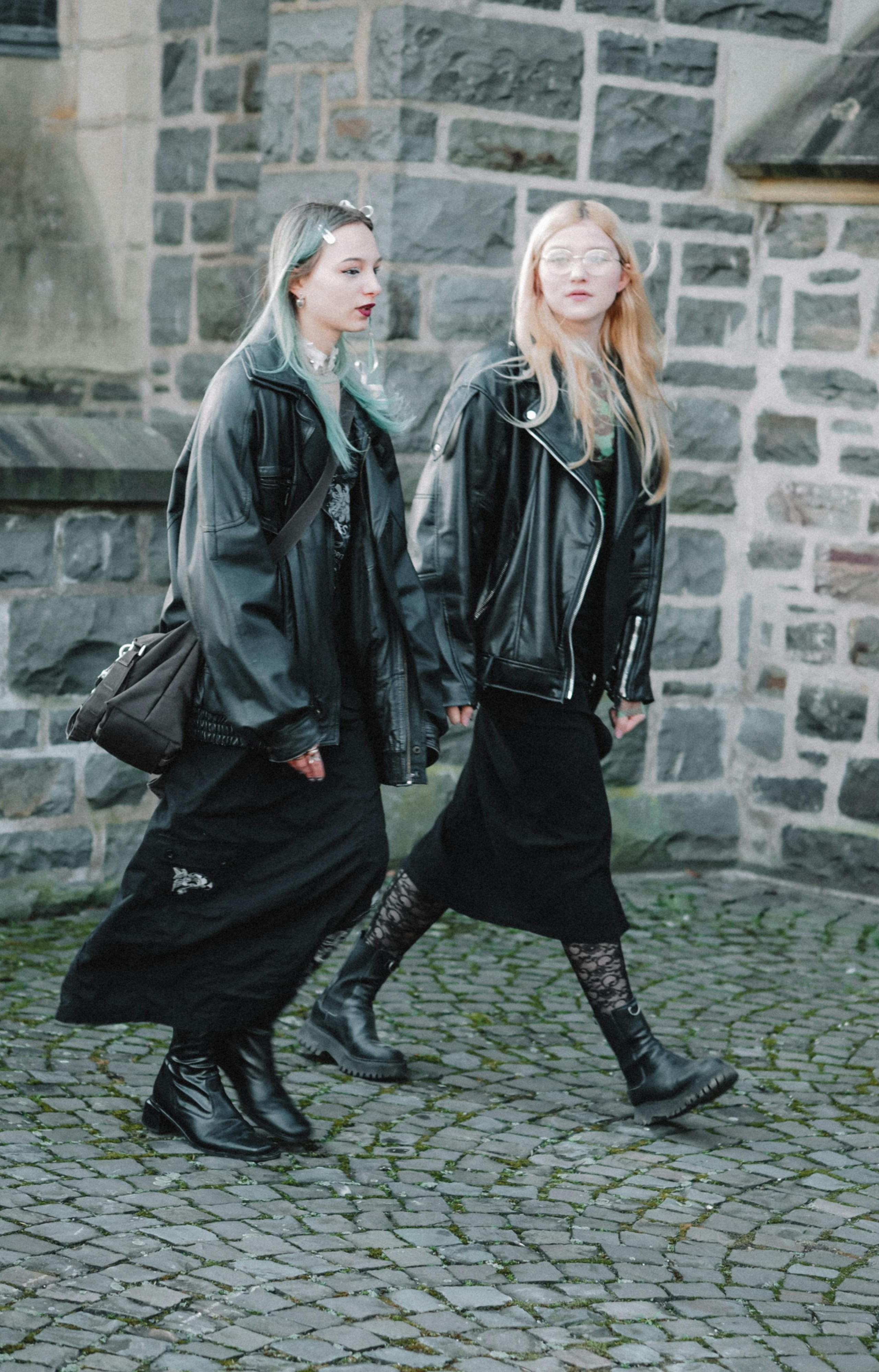 Two people wearing long black outfits with leather jackets, black boots, and lace or patterned tights.