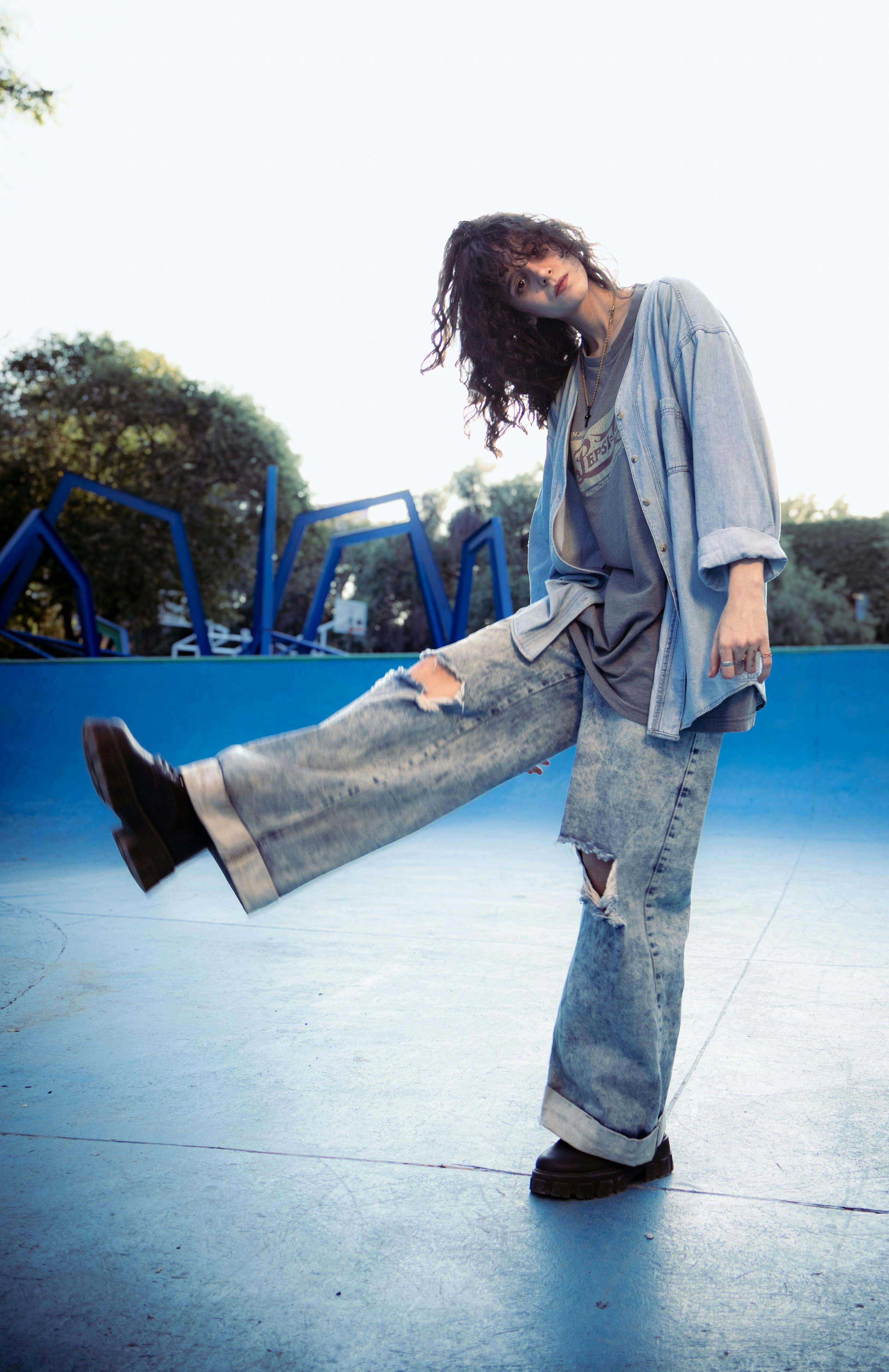 Person wearing a light denim shirt over a graphic T-shirt, loose ripped jeans, and black shoes at a skate park.
