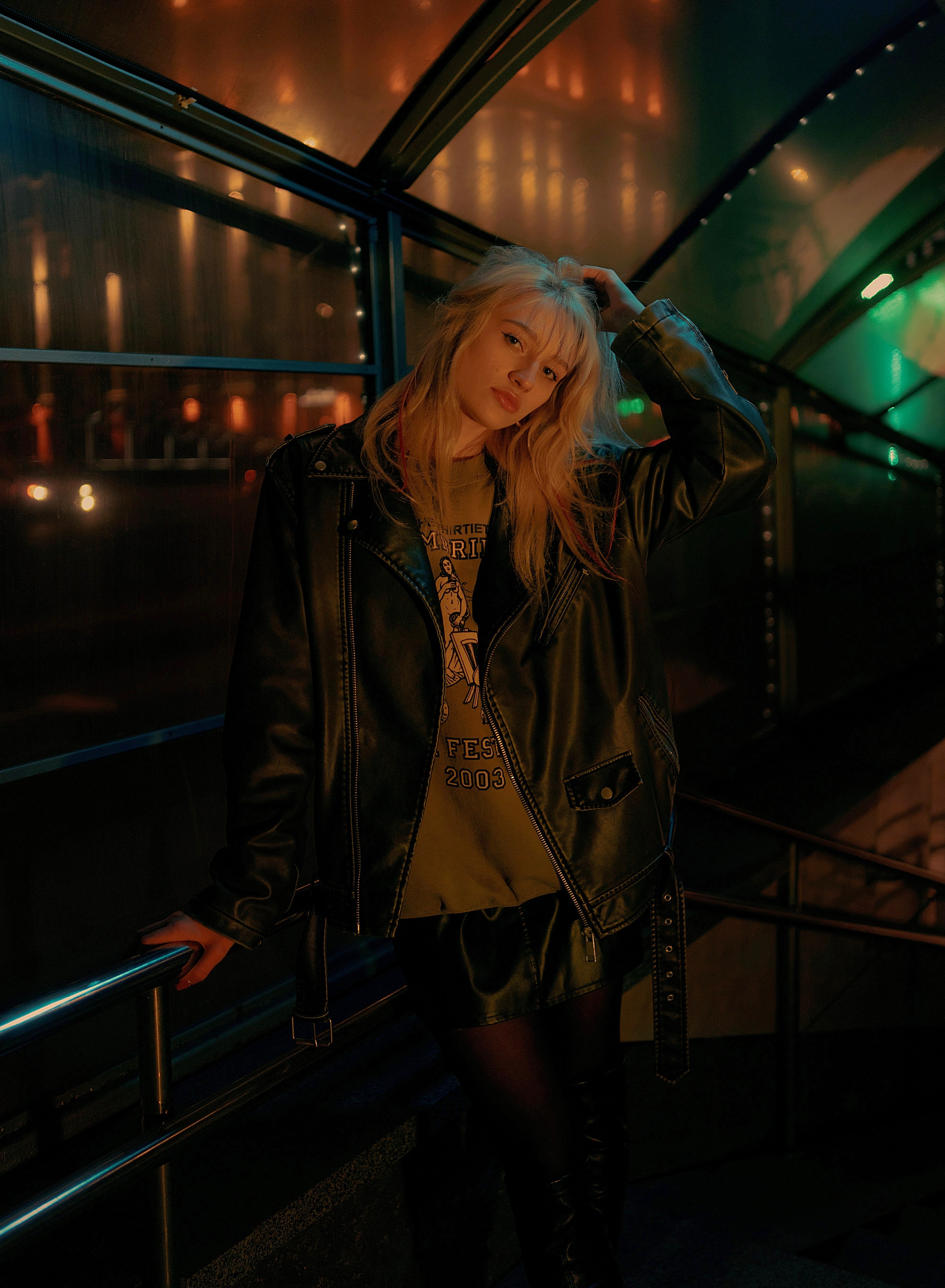 Person wearing a black leather jacket over a graphic T-shirt, black shorts, tights, and tall boots in a dimly lit walkway.