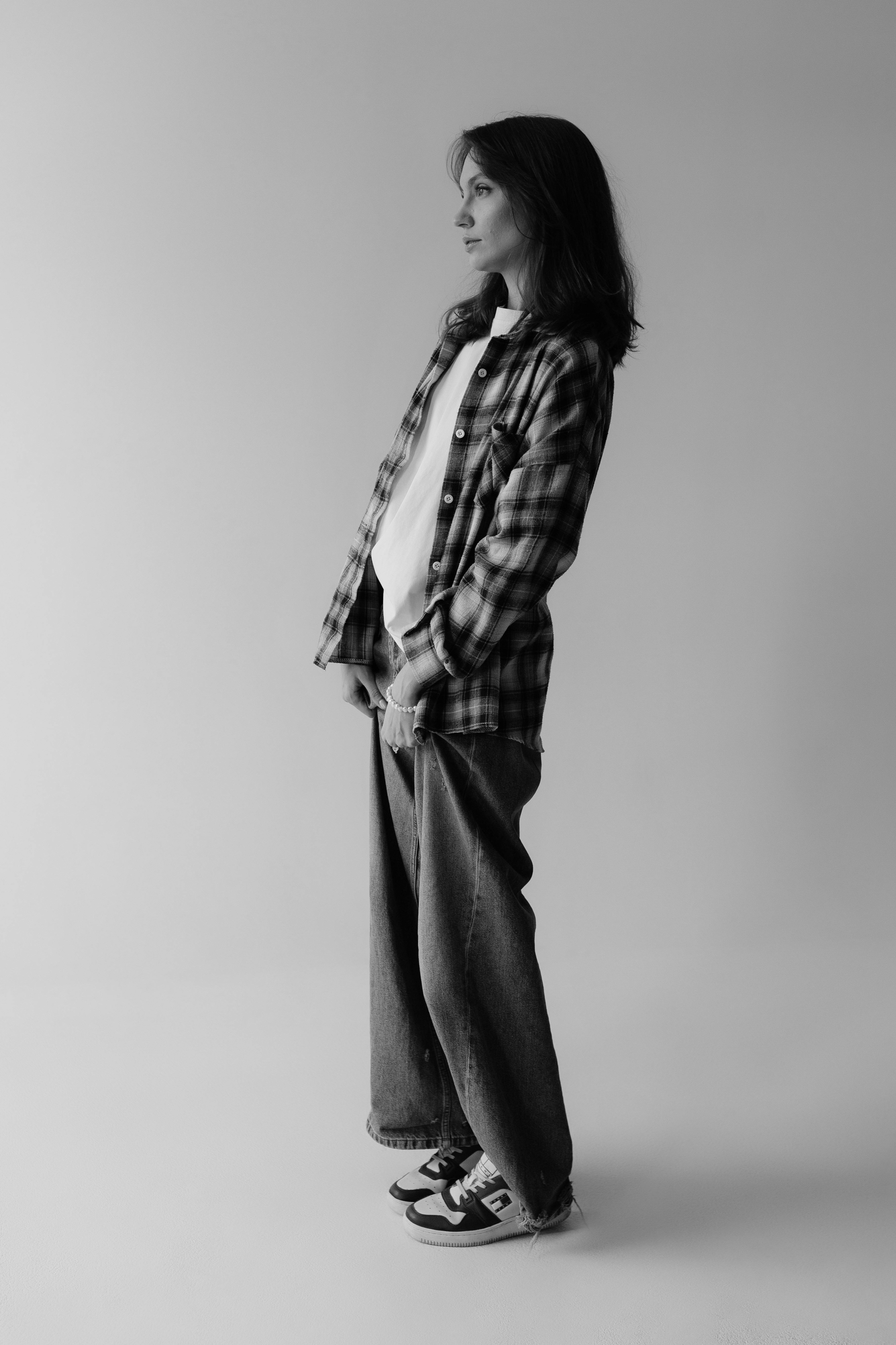 Black and white image of a person wearing a plaid flannel over a white shirt, loose jeans, and sneakers.