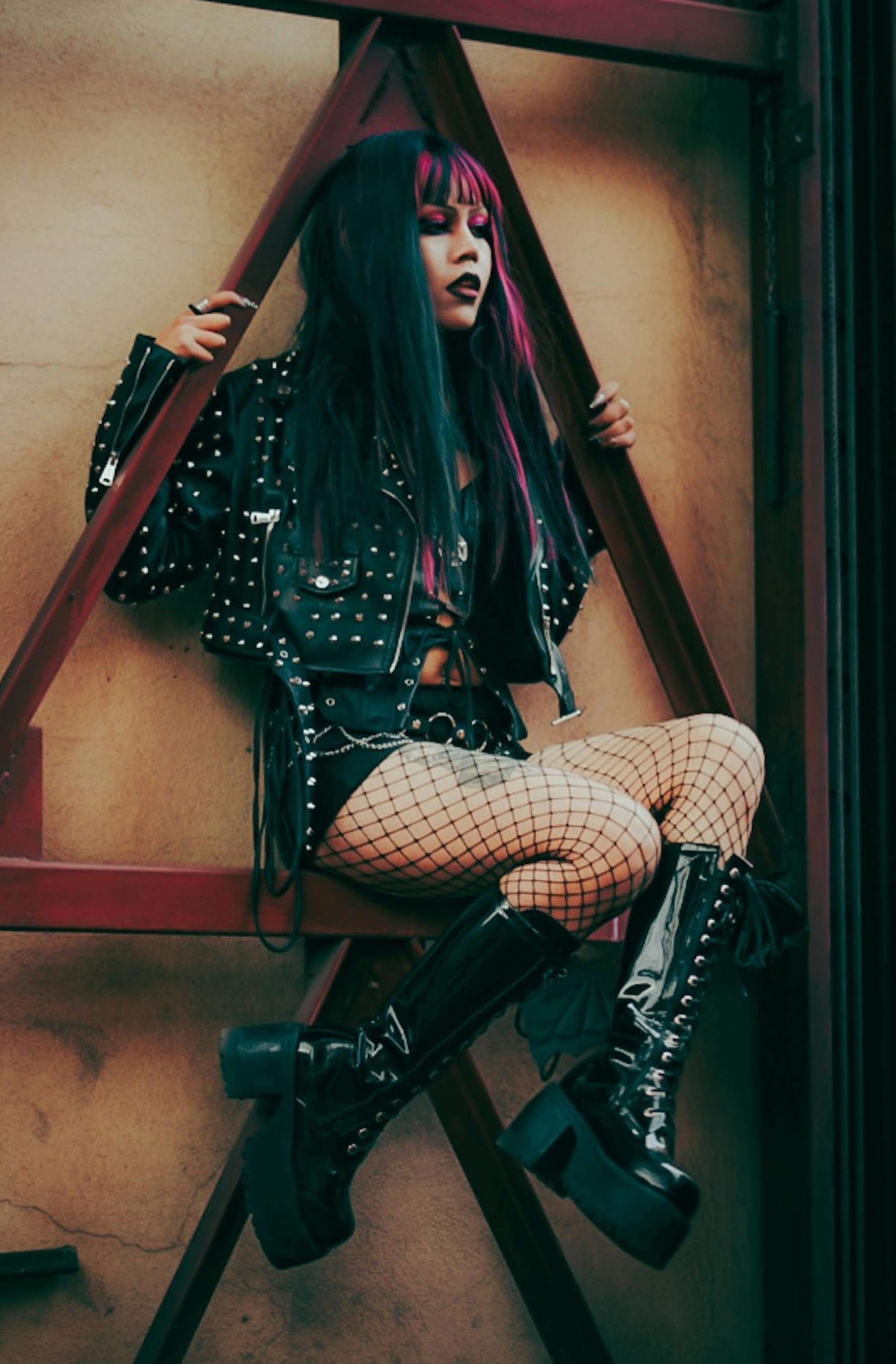 Person wearing a studded black jacket, black outfit, fishnet tights, and tall glossy platform boots while sitting on a metal structure.
