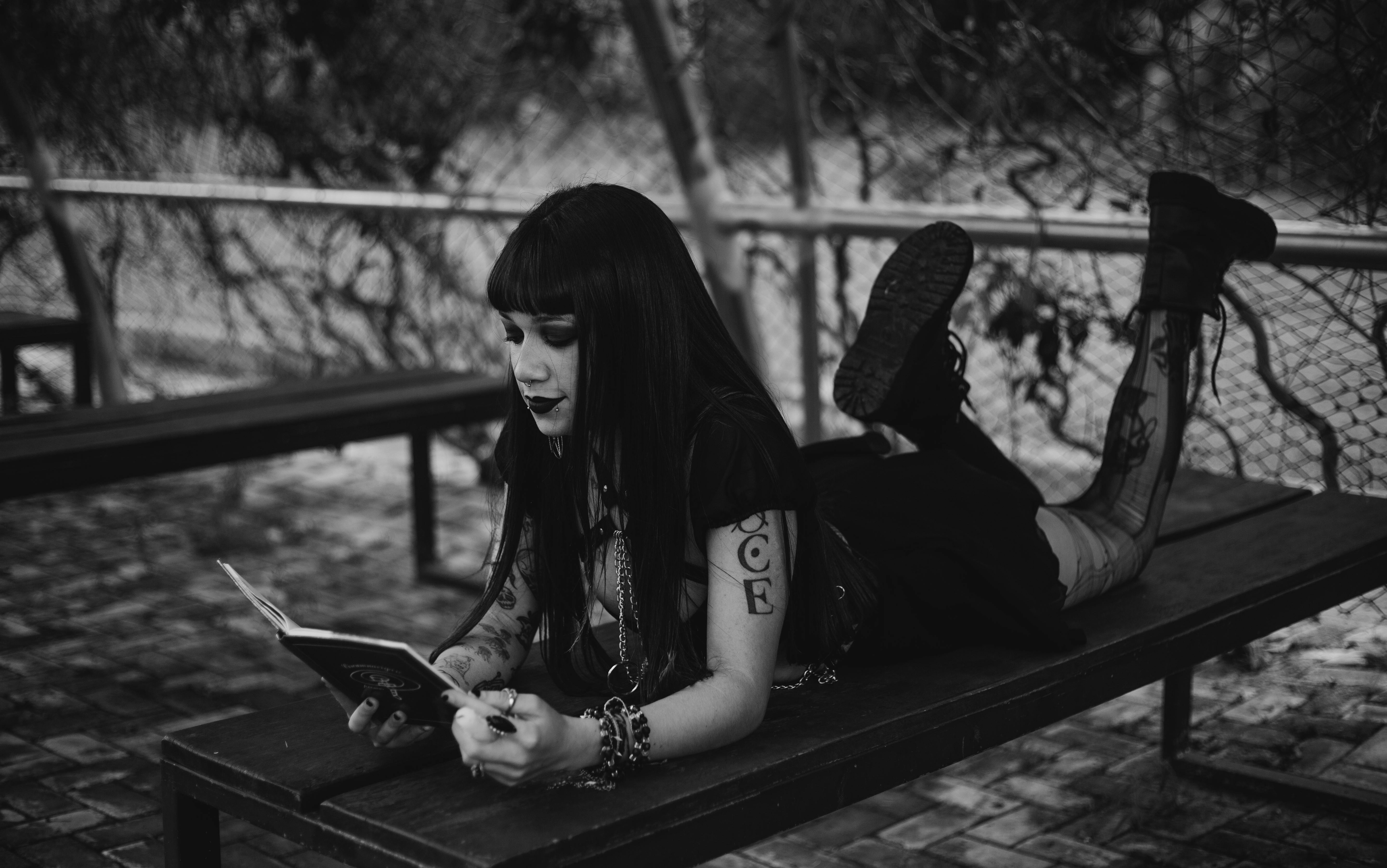 Black and white image of a person lying on a bench while reading, wearing a black outfit with chains, bracelets, and tall boots.