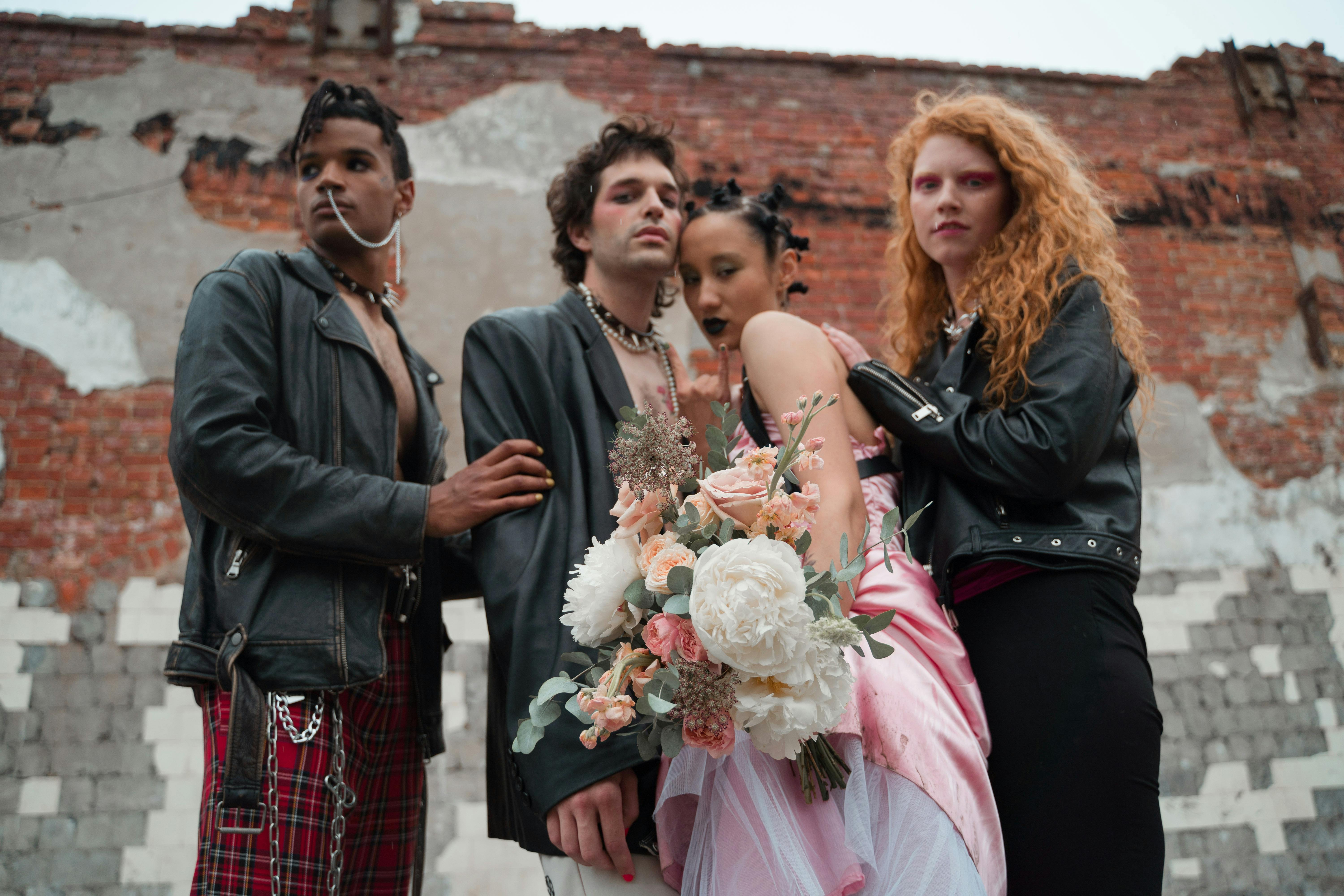 Group of four people wearing alternative outfits with leather jackets, chains, red plaid pants, a pink dress, and a bouquet of flowers.