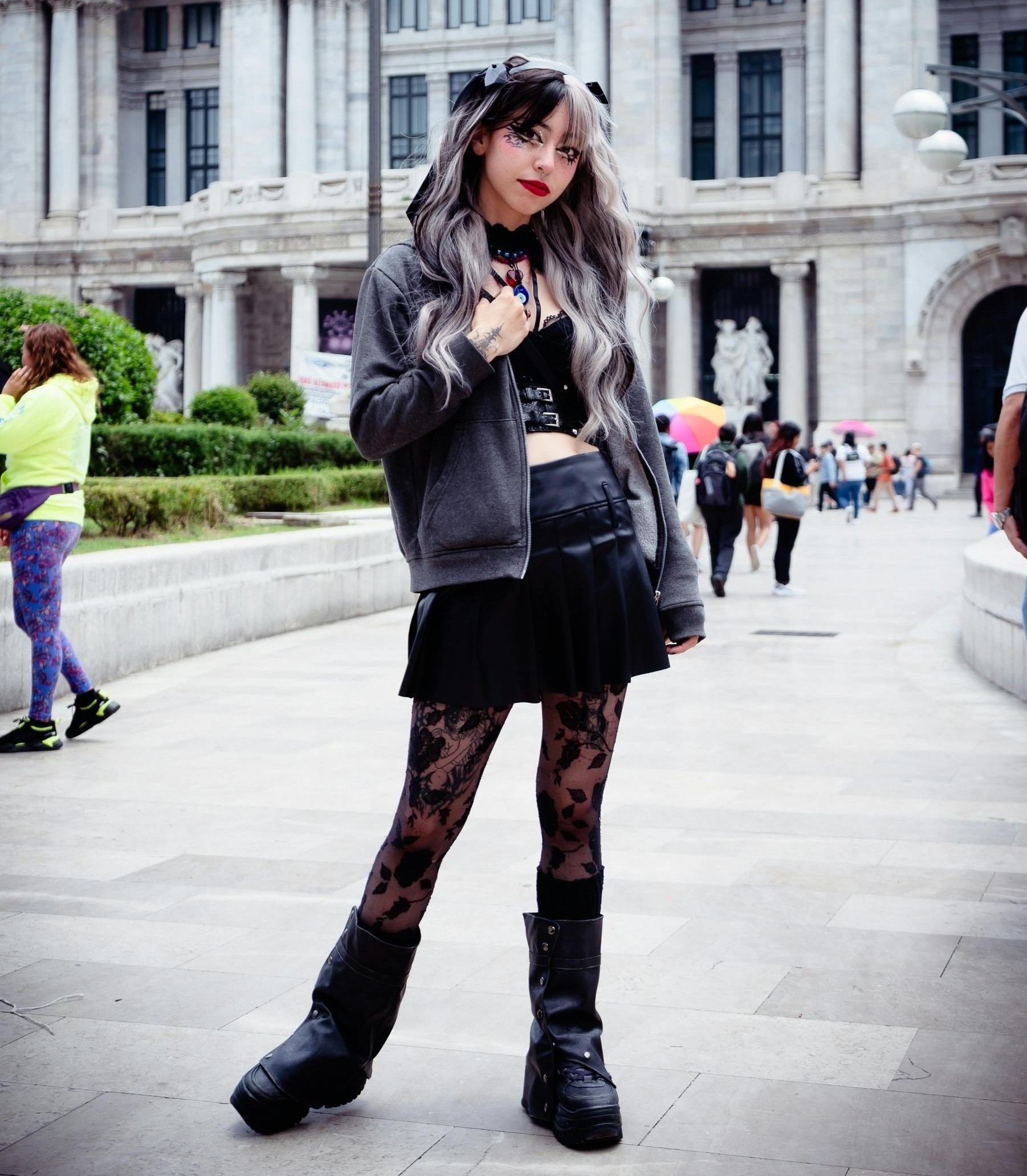 Person wearing a gray hoodie, black skirt, lace tights, and tall black boots in front of a large building.