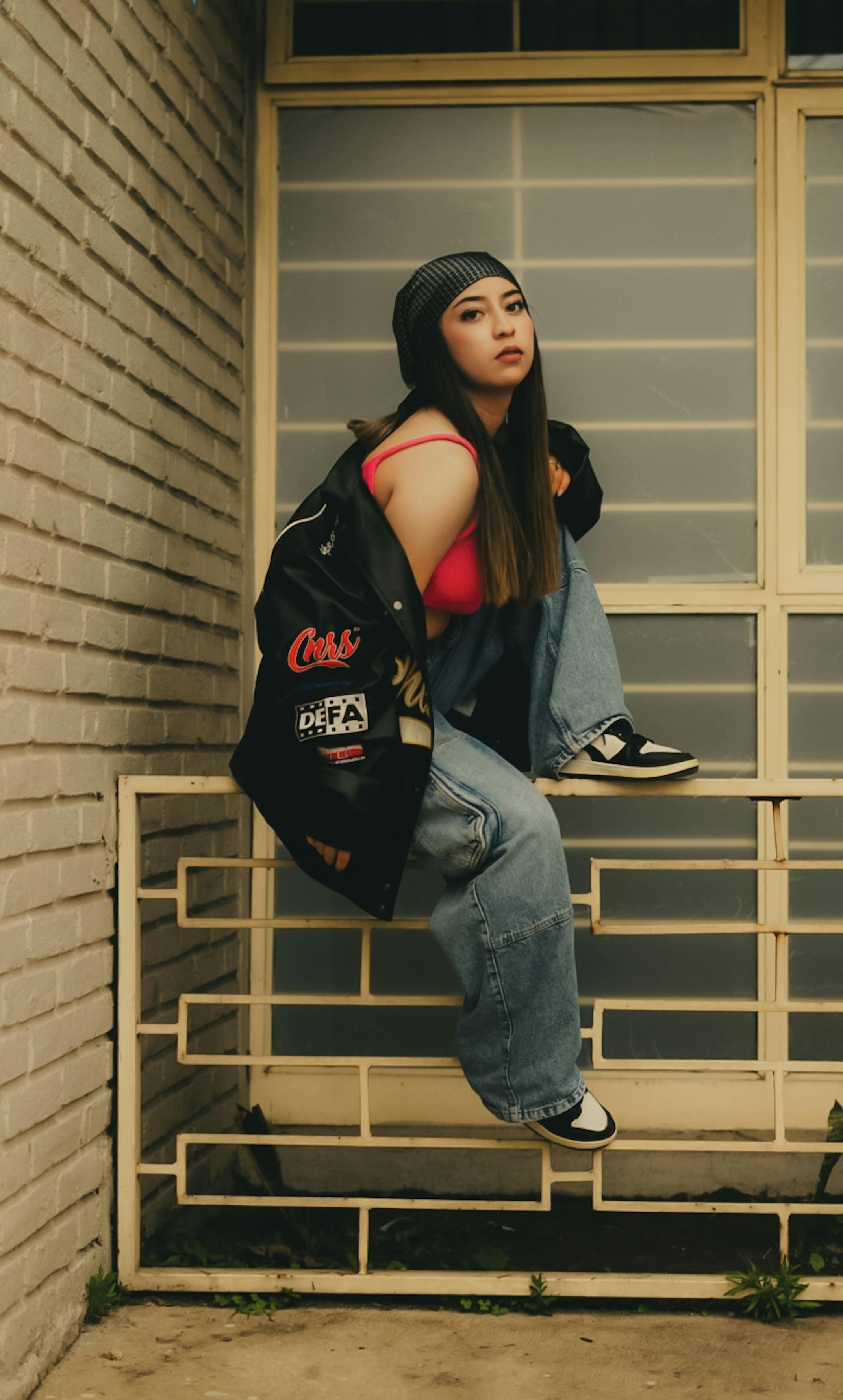 Person sitting on a railing wearing a black and white jacket, jeans, sneakers, and a beanie.