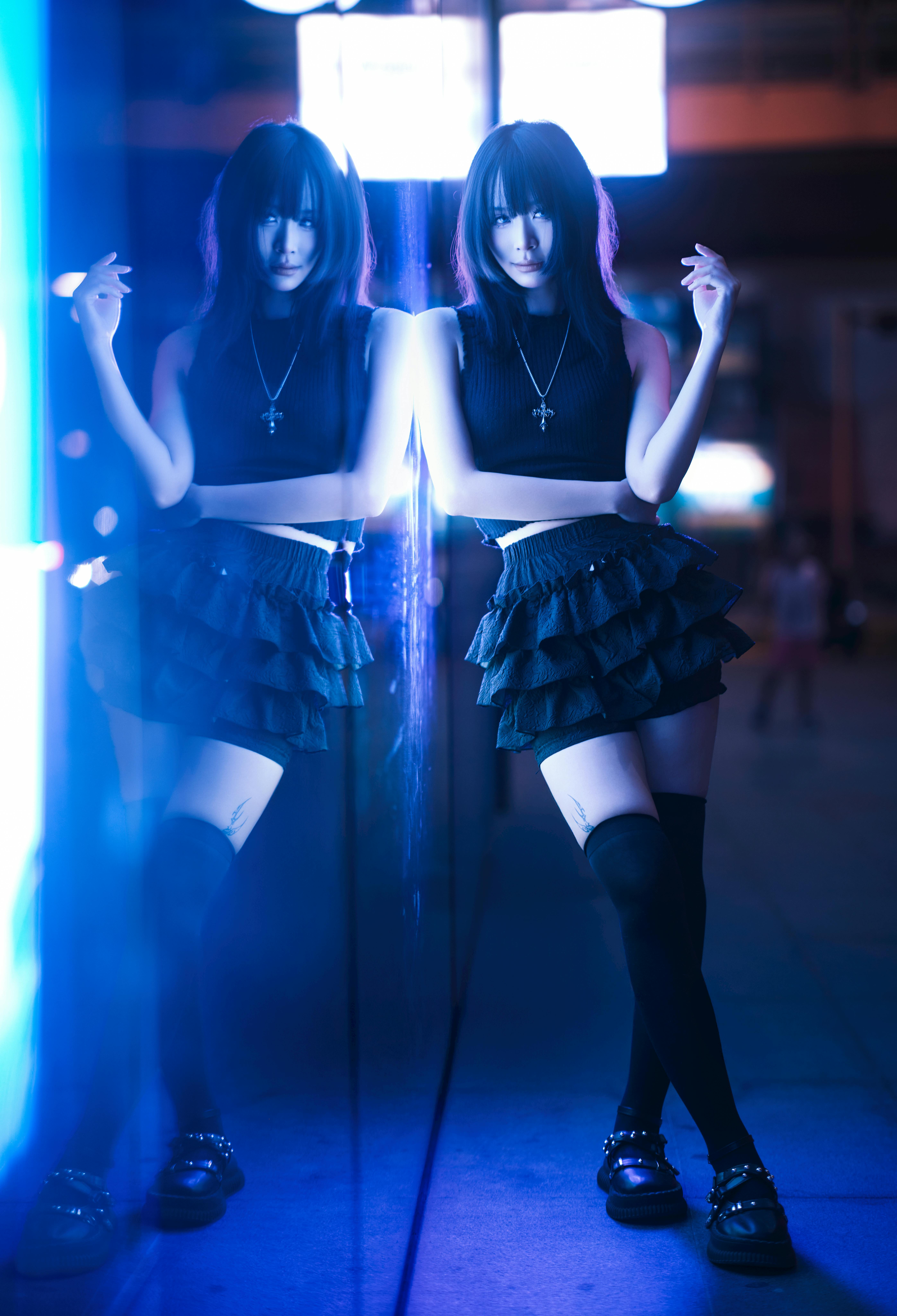 Person standing near a mirror in blue lighting wearing a black sleeveless top, ruffled black skirt, tall socks, and black shoes.