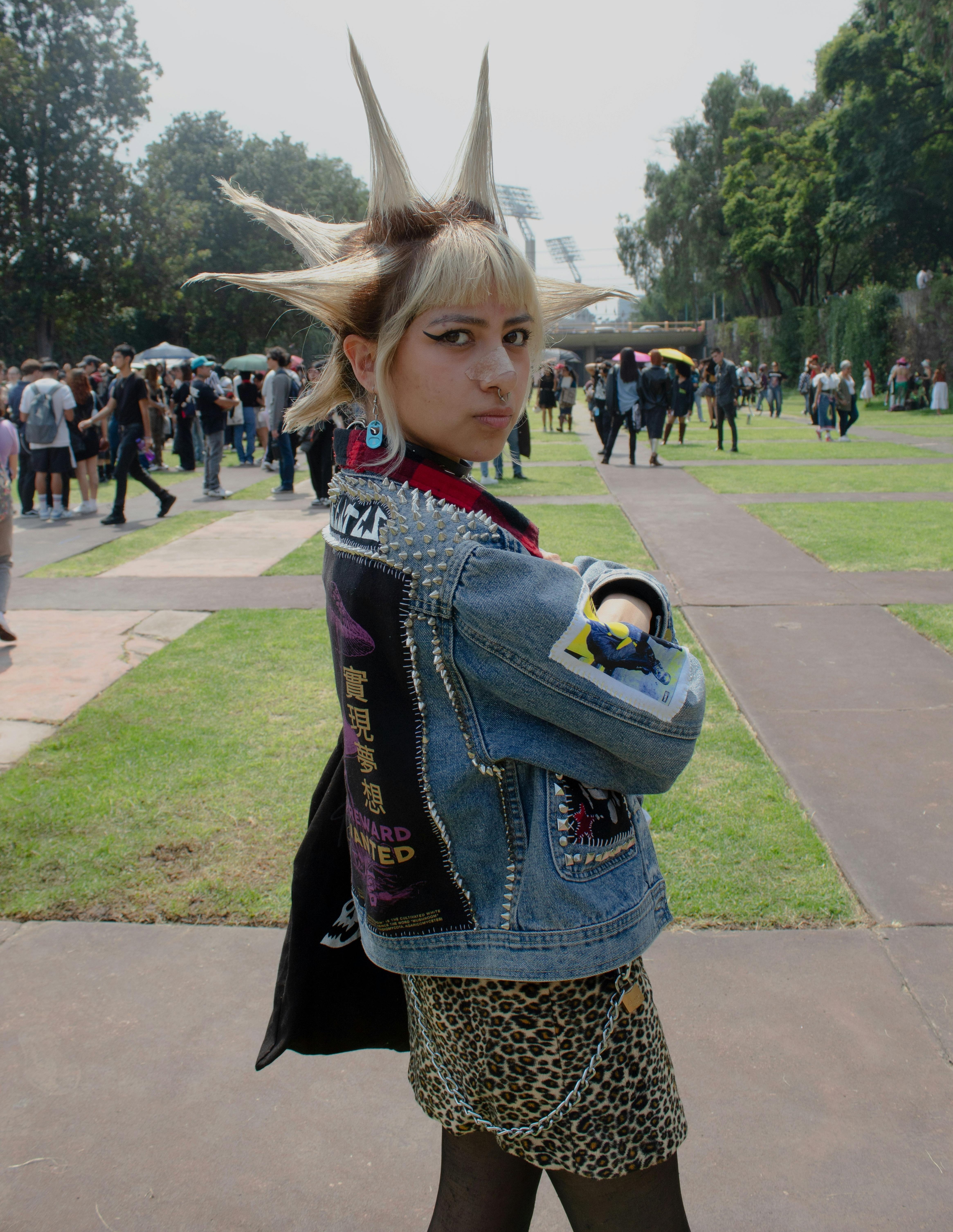 Person with spiked blonde hair wearing a studded denim jacket, leopard print skirt, and chain details at an outdoor event.