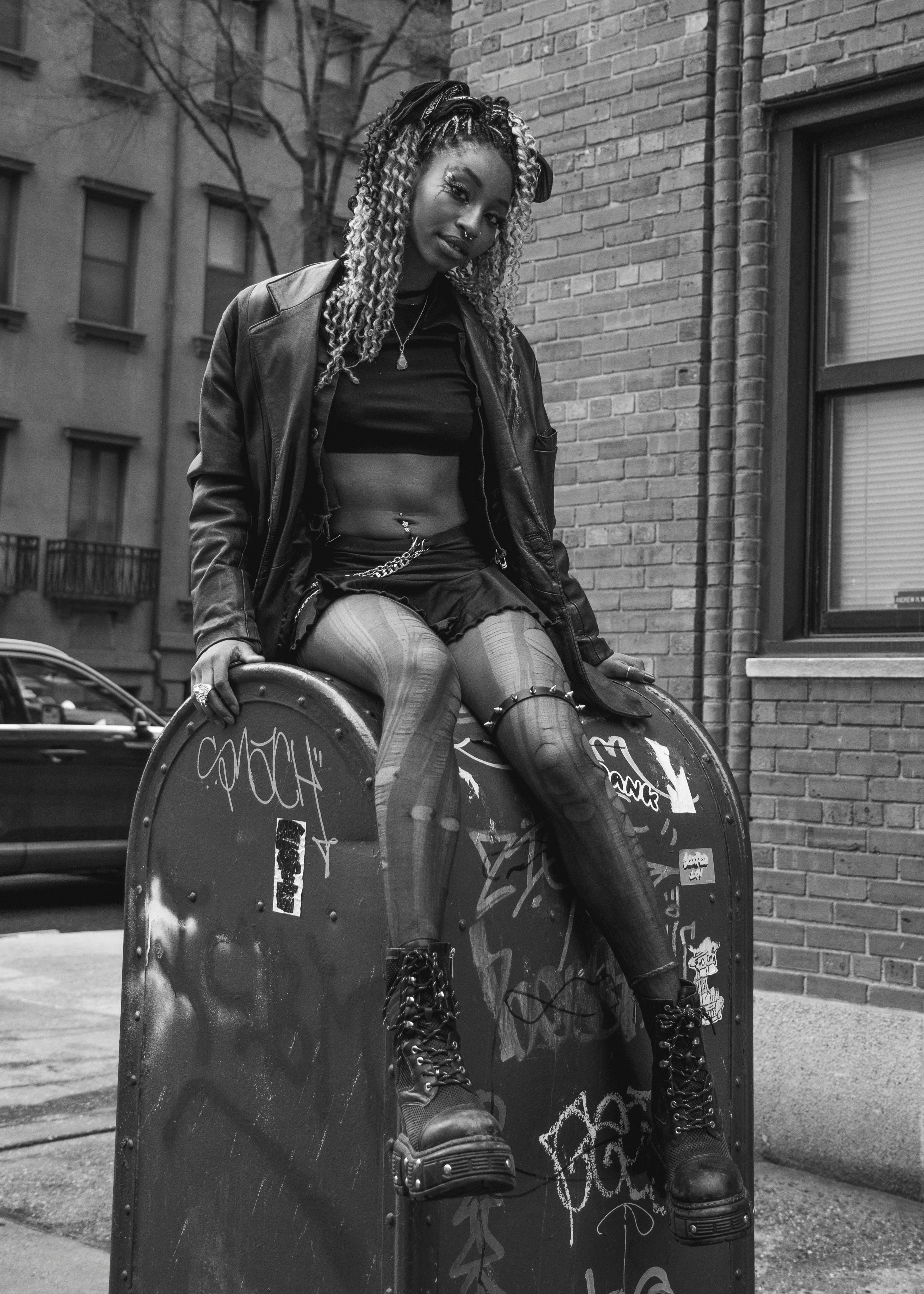 Black and white image of a person sitting on a graffiti-covered mailbox wearing a leather jacket, black skirt, patterned tights, and lace-up boots.