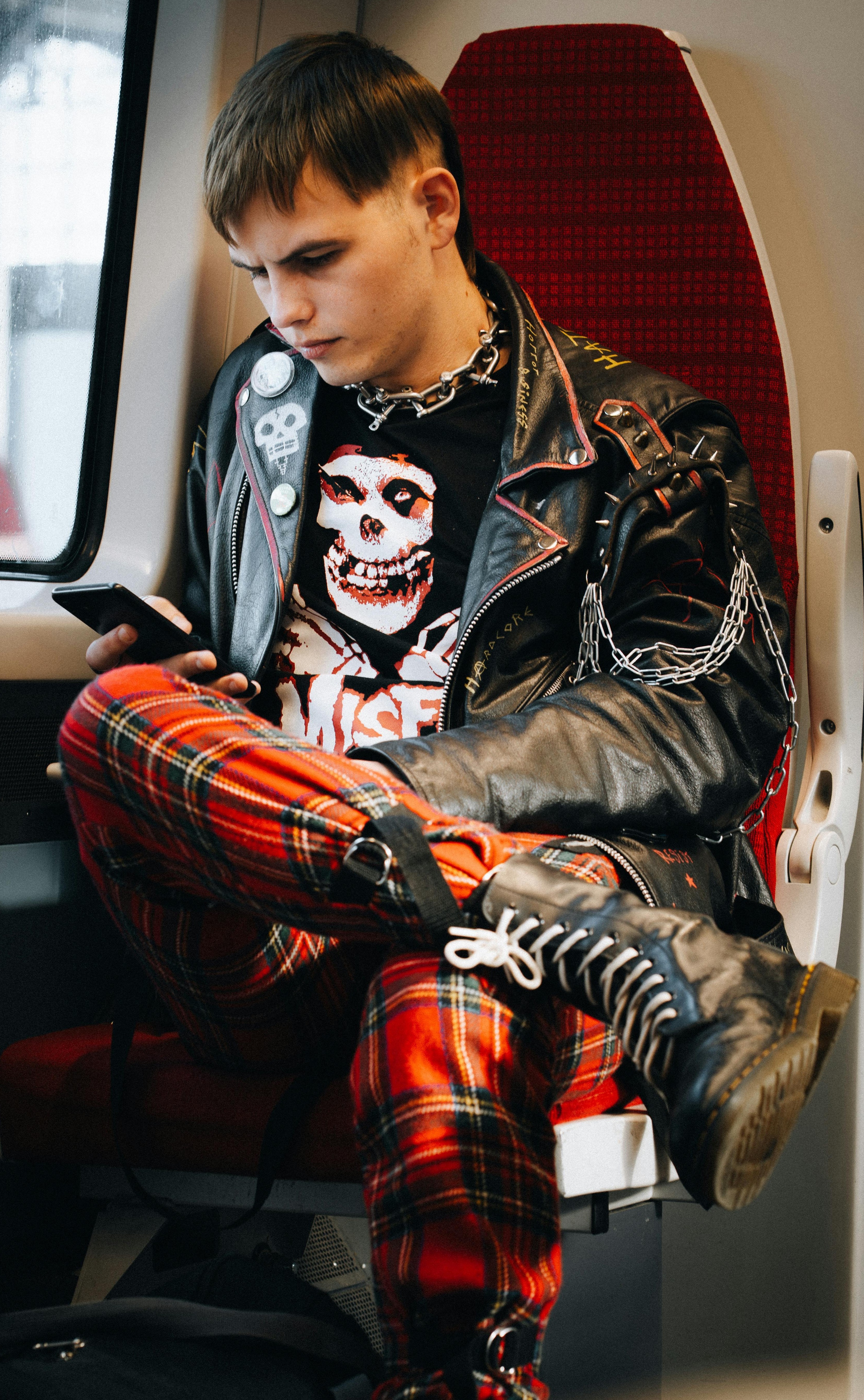 Person sitting on a train wearing a black leather jacket with spikes and chains, red plaid pants, black boots, and a skull graphic shirt.