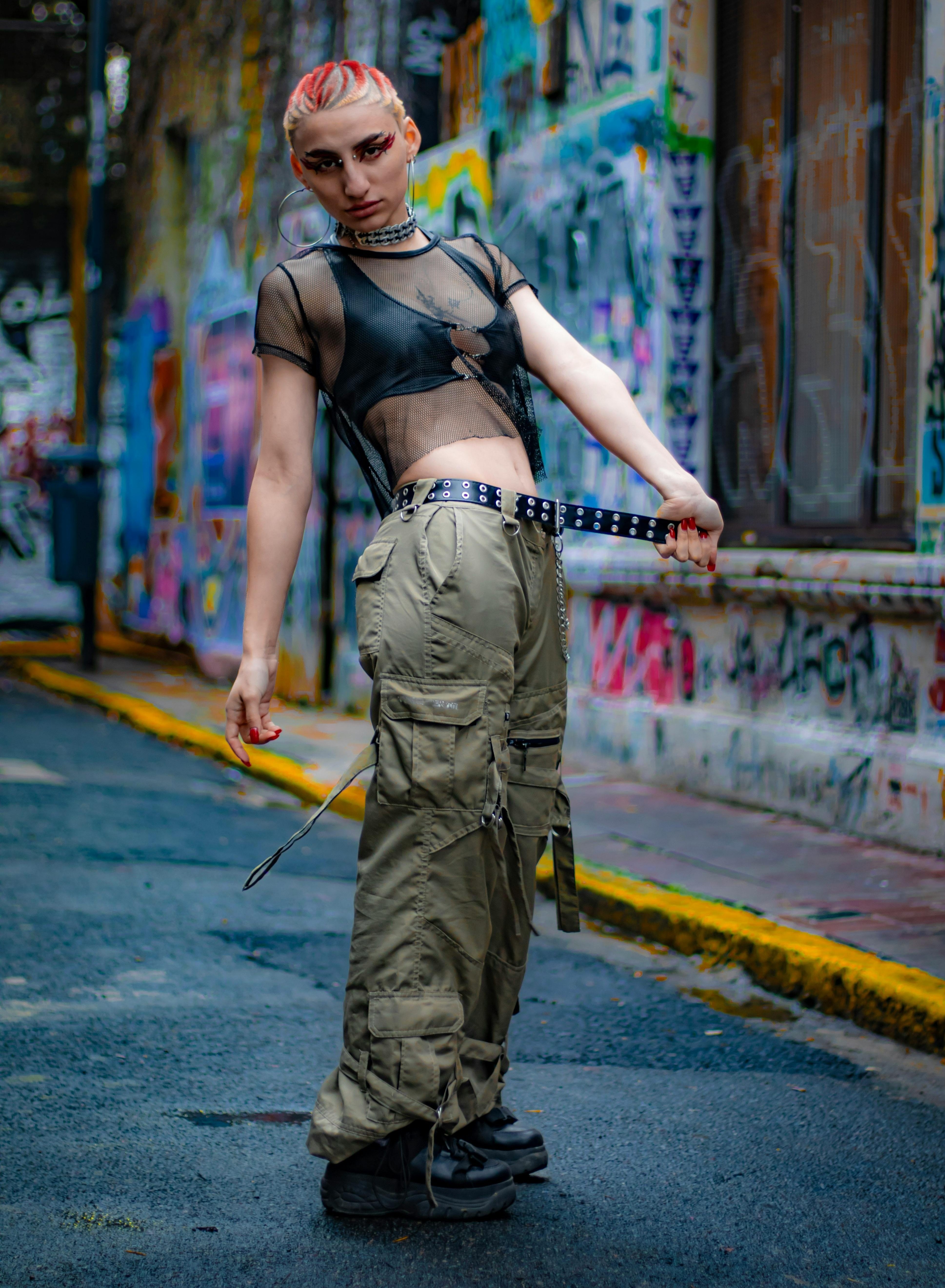 Person wearing a sheer black top, khaki cargo pants, studded belt, chain necklace, and black shoes in front of a graffiti-covered wall.